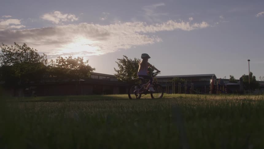 The video shows a wide shot of a grassy field with a building and playground in the background a child wearing a helmet rides a bicycle from the left side of the frame going past the camera and into the distance