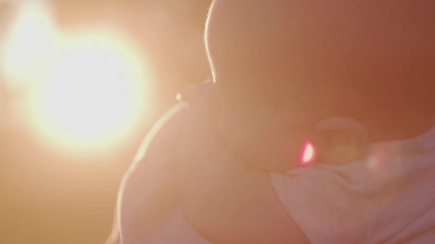 The back of a babys head and shoulder as they are being held affectionately by an adult with a bright warm light creating a soft glow behind them