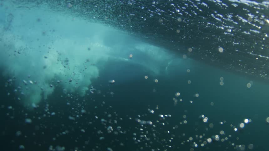 An underwater view shows numerous bubbles of varying sizes rising in blue water as sunlight streams in from above