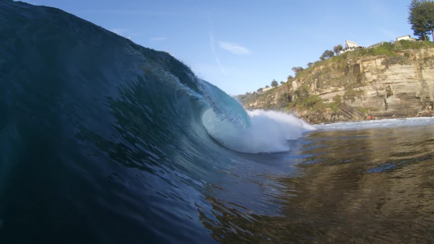 Houses sit on a seaside cliff as a large breaking wave flows by covering the area near the base of the cliff in water
