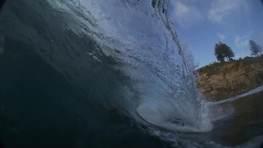 A dynamic lowangle shot captures a powerful blue wave curling and breaking over a rocky shoreline under a partly cloudy sky