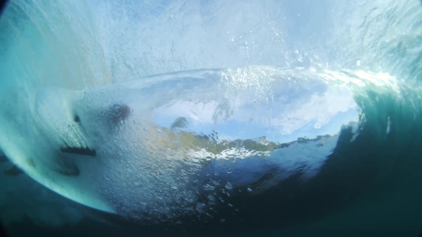 The video shows a unique perspective from underwater looking up at the surface of the sea as a wave curls and breaks the bright blue sky and clouds are visible through the shimmering turbulent water and a surfer can be seen riding the wave from below