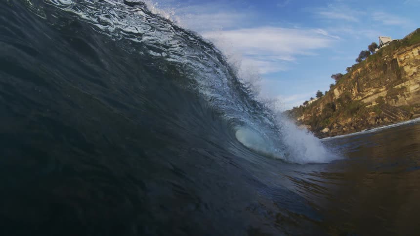 A powerful wave curls near the rocky coastline under a clear blue sky