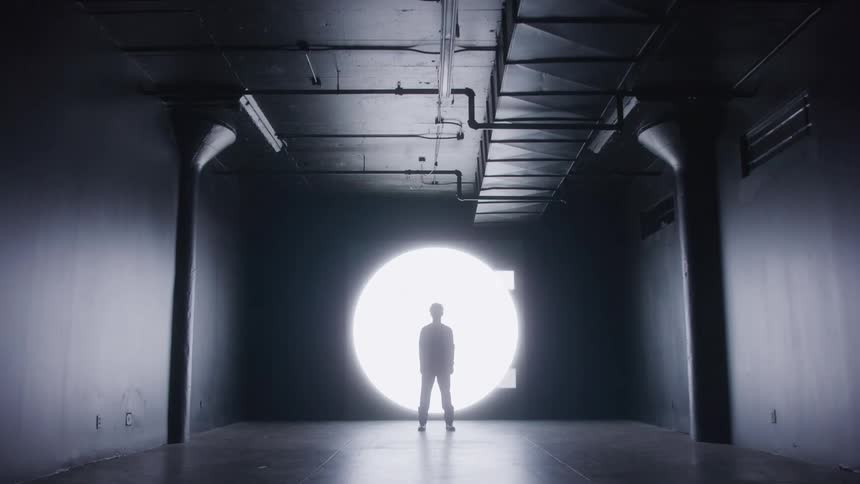 A single person is silhouetted in the center of a large circular white light against a dark wall while several small square lights surround them