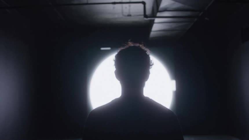 A silhouetted figure walks down a dark hallway towards a bright circular light at the end