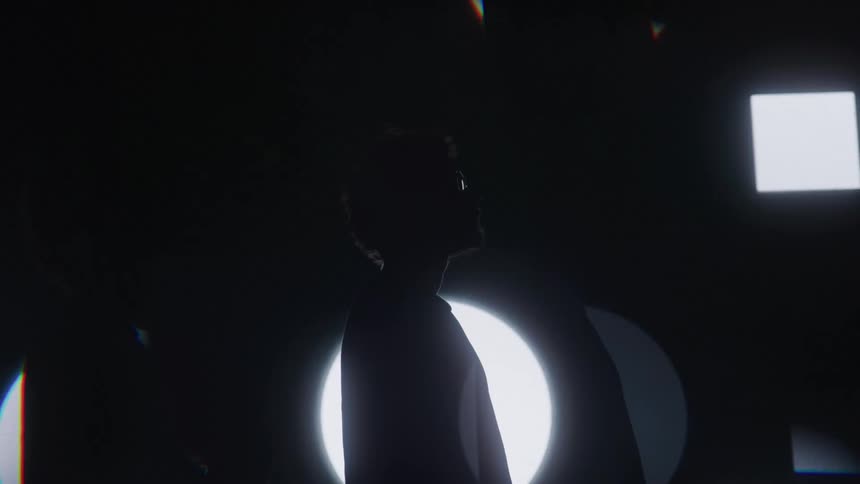 The video shows a man standing in a dark room he is backlit by a large circular light that pulses in a hypnotic way smaller square lights arranged around him also turn on and off in a pattern