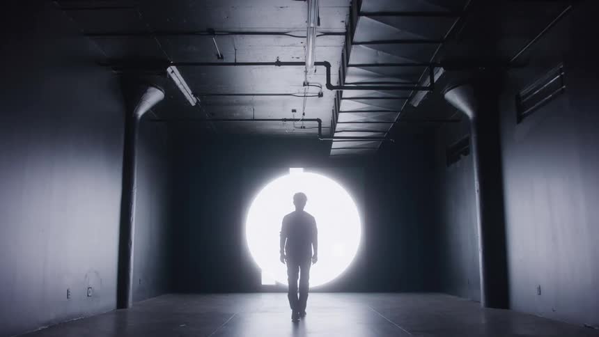 Silhouette of the back of a man in a dark room walking towards flickering lights on the wall indoors