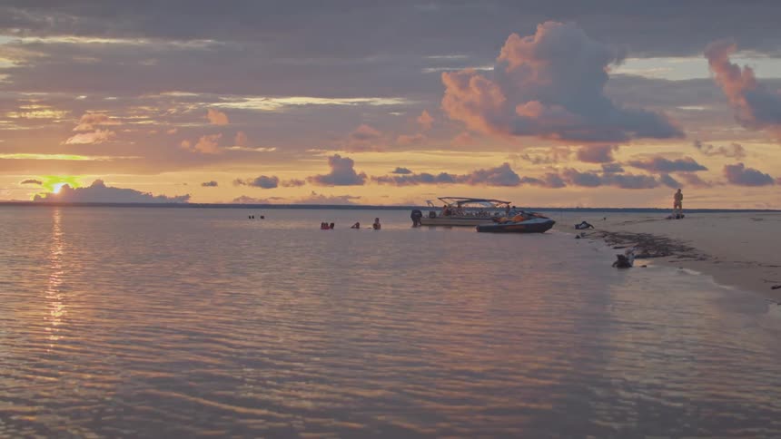 The video shows a beautiful sunset over the water with a small boat and some people near the shore the sky is filled with soft clouds in shades of orange pink and purple