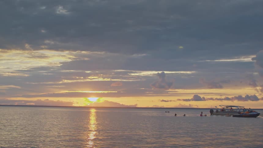 The sun dips over a calm ocean a small boat and distant swimmers silhouetted against glowing clouds