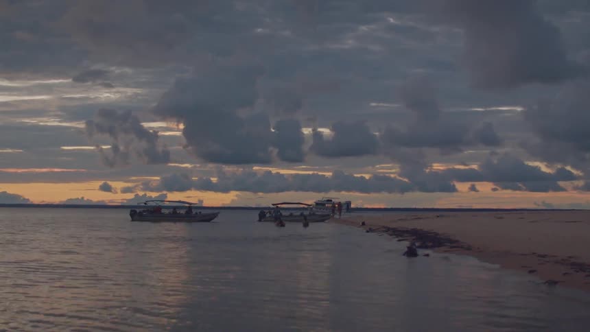 The video shows a wide shot of a river or lake at sunset with a person paddling a kayak near the shore there are also a few small boats anchored nearby the sky is full of dramatic dark clouds with a streak of orange light on the horizon