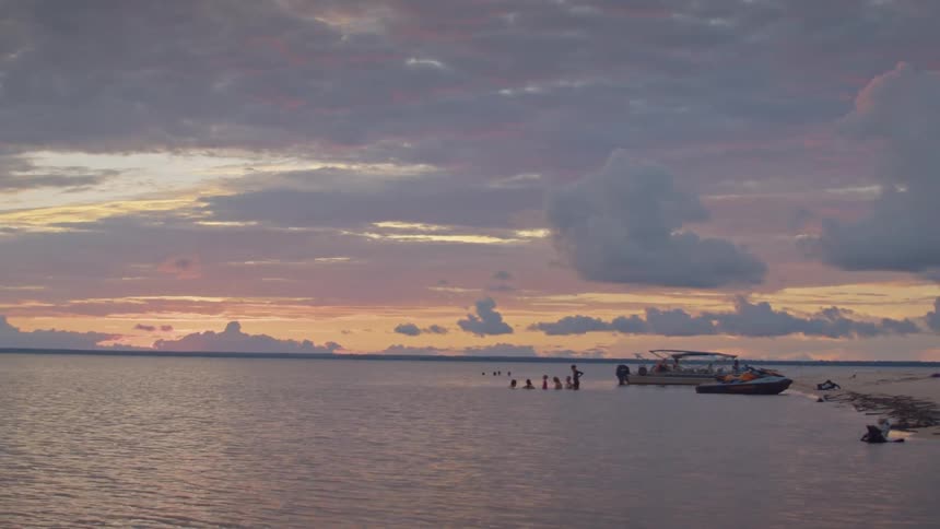 Boats anchored near people in the water beside a sandbar at sunset