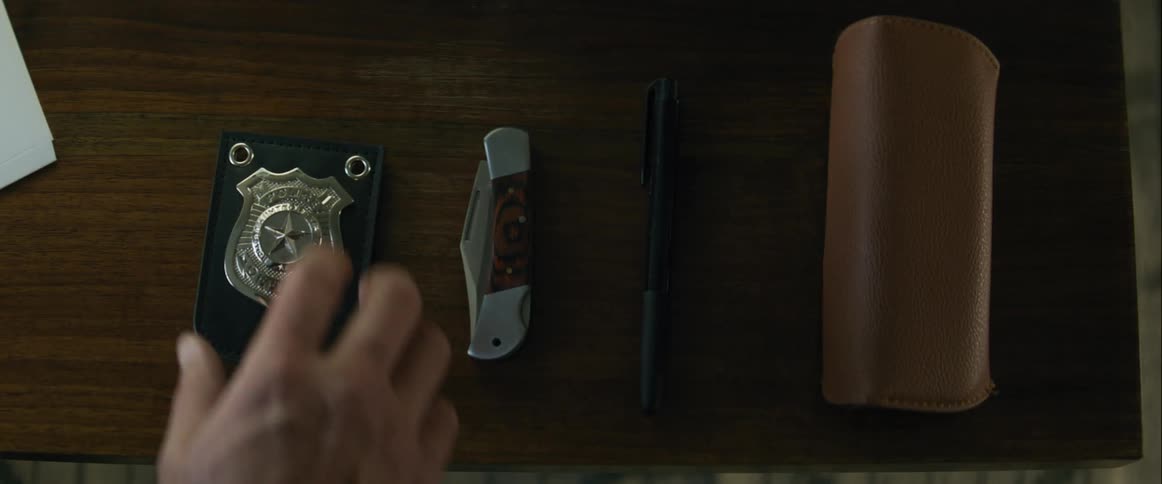Hand reaches for a badge on a table next to a pocketknife pen and brown case
