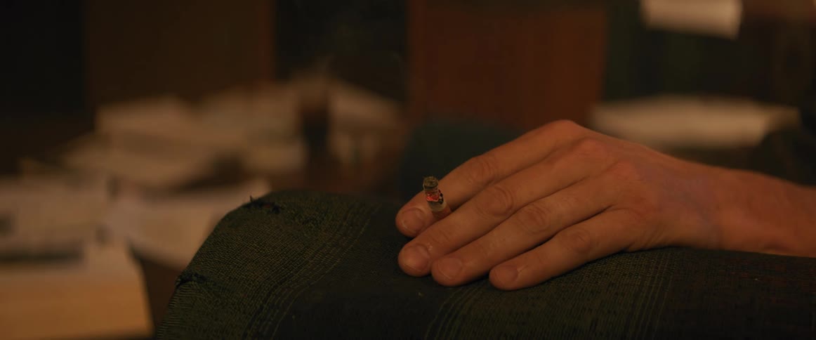 Hand holds a lit cigarette resting on the arm of a dark chair