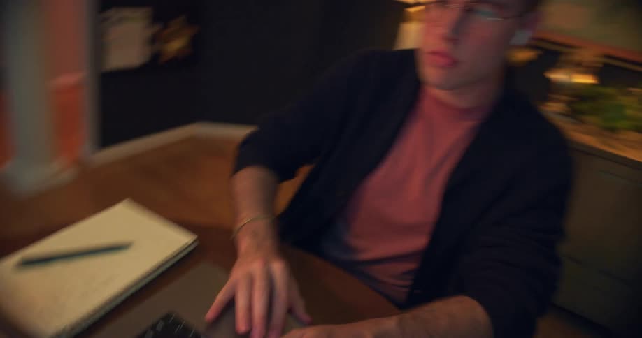 White man wearing glasses sitting down and typing on a laptop computer indoors