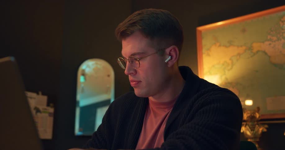 A young man wearing glasses and earbuds sits on a laptop in a partially dark room