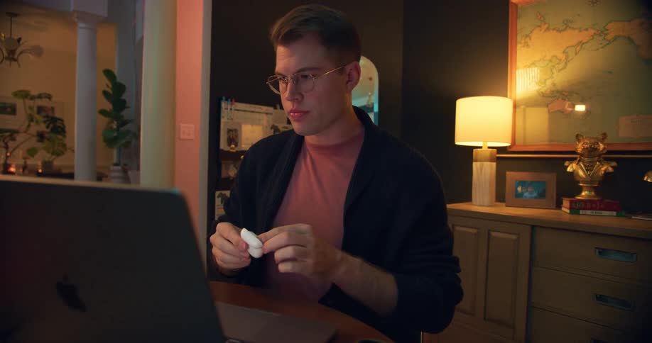 The video shows a man with short reddishblonde hair and glasses sitting at a desk hes wearing a dark cardigan and puts on a pair of wireless earbuds before looking at the laptop screen in front of him