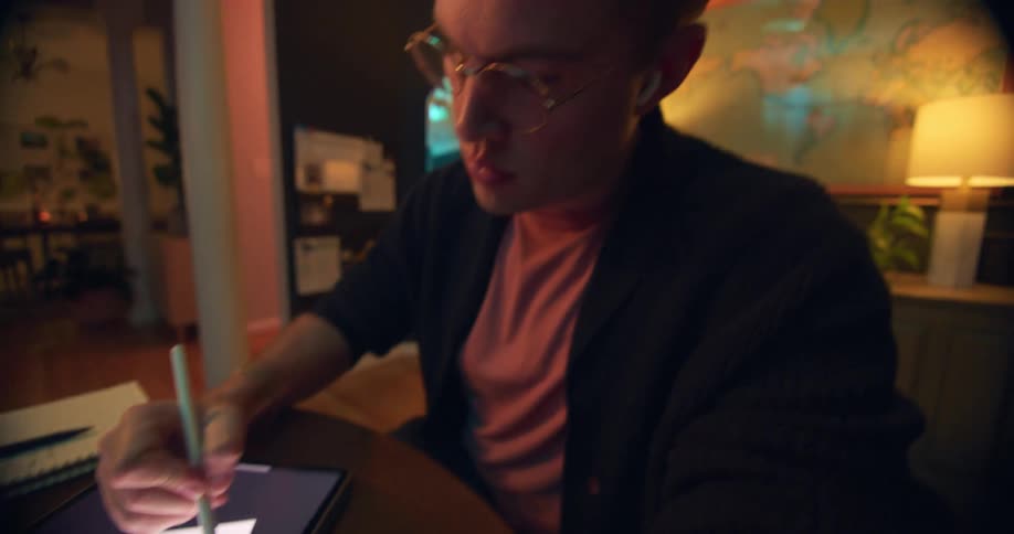Young man wearing glasses sitting down at a table in a living room drawing on a tablet indoors