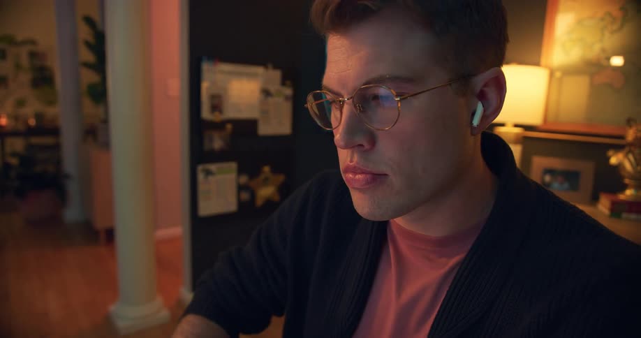 The video shows an overhead shot of a man sitting at a desk with a laptop and a notebook he is wearing wireless earbuds and working on his computer he closes the laptop and the camera shifts to a closeup of his face as he looks thoughtfully into t