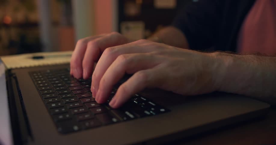 A close view shows a persons hands typing on a black laptop keyboard in a dimly lit room