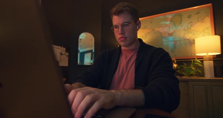 A young man types and uses a laptop computer while sitting at a table indoors