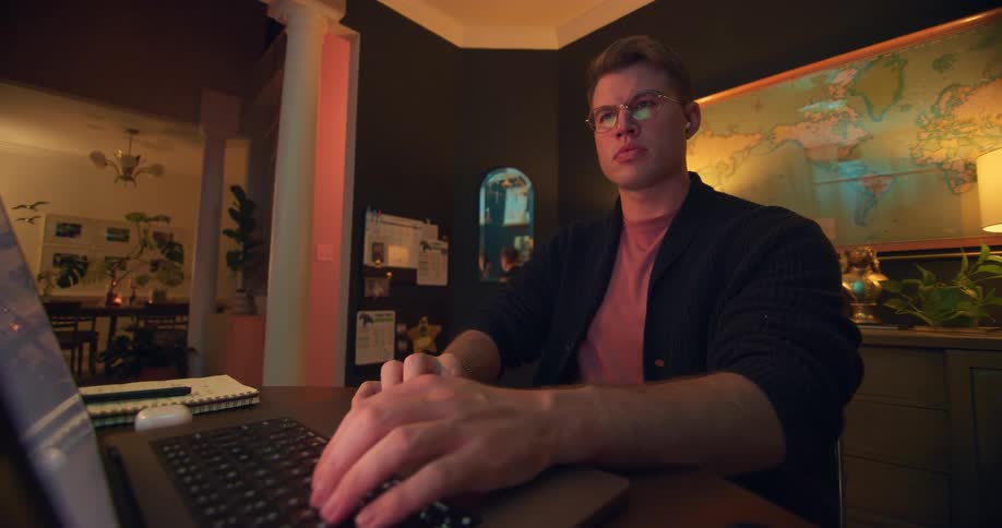 A young man wearing glasses sits at a desk in his home office typing on a laptop
