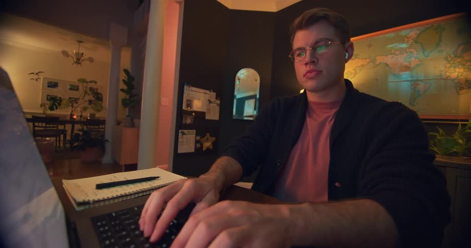 A young man wearing glasses opens his macbook and types on it in a home office