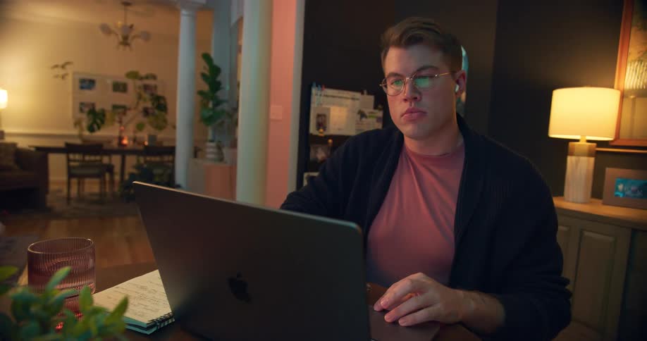A man with glasses is sitting at a desk and working intently on his laptop with a glass of water a notepad and a lamp also on the table