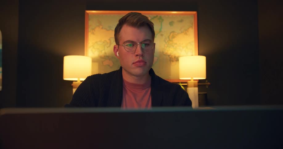 White man wearing glasses opening the lid of a laptop computer and staring at the screen indoors