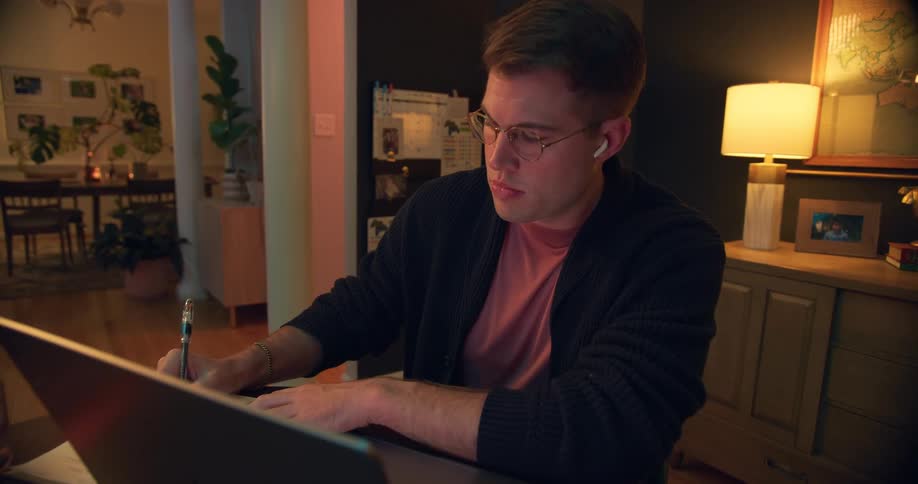 Man wearing glasses and earbuds writes in a notebook while working in front of a laptop