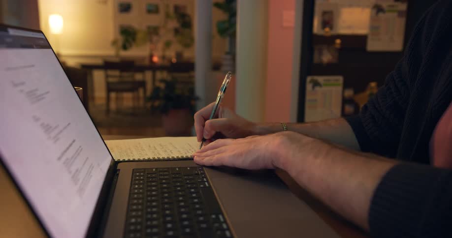 Young adult man wearing glasses sitting down indoors writing with a pen on a notebook while looking at a laptop computer screen