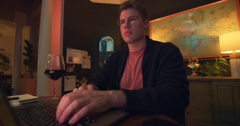 A man works on his laptop at a desk with a glass of wine by his side