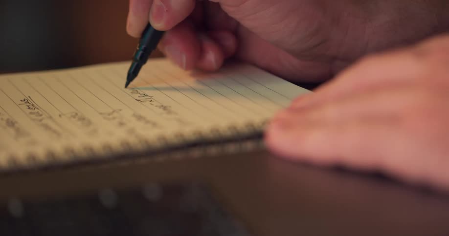 White male hands writing on a notebook using a pen next to a computer keyboard indoors