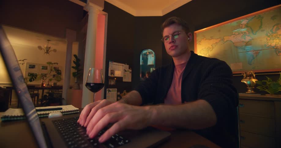A focused man in glasses is sitting at a desk and typing on his laptop he has a glass of wine on the desk and a world map on the wall behind him