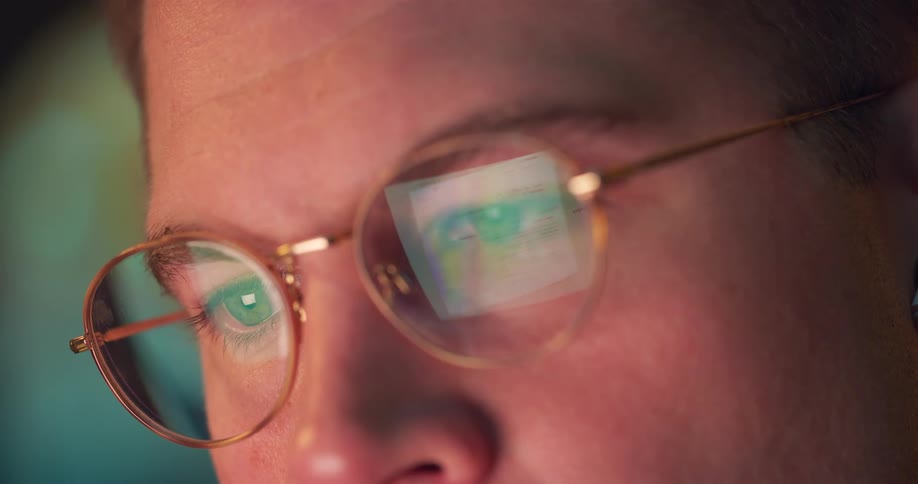 A closeup shot of a persons face with a focus on their eyes and the reflection of a computer screen on their round glasses