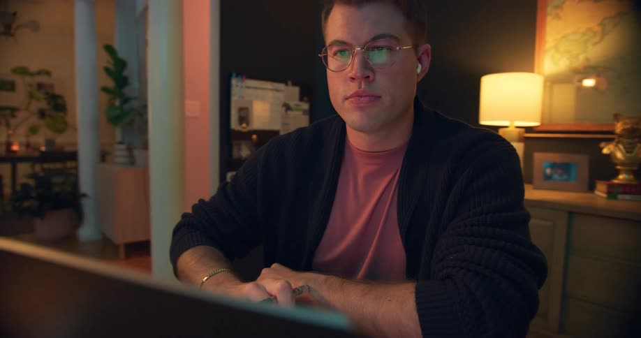 Wearing earbuds a man concentrates at his laptop in a cozy workspace lit by a warm desk lamp