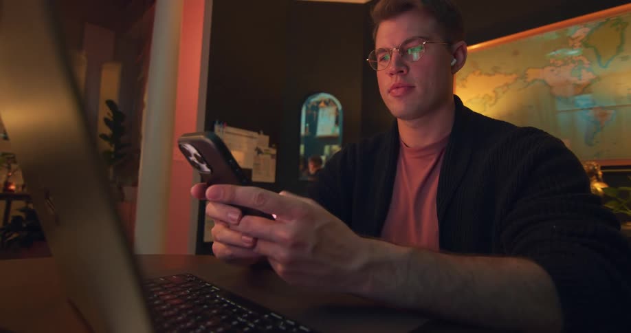 White man wearing glasses sitting down and typing on a smartphone while looking at a laptop screen on the table in front of him indoors during day time