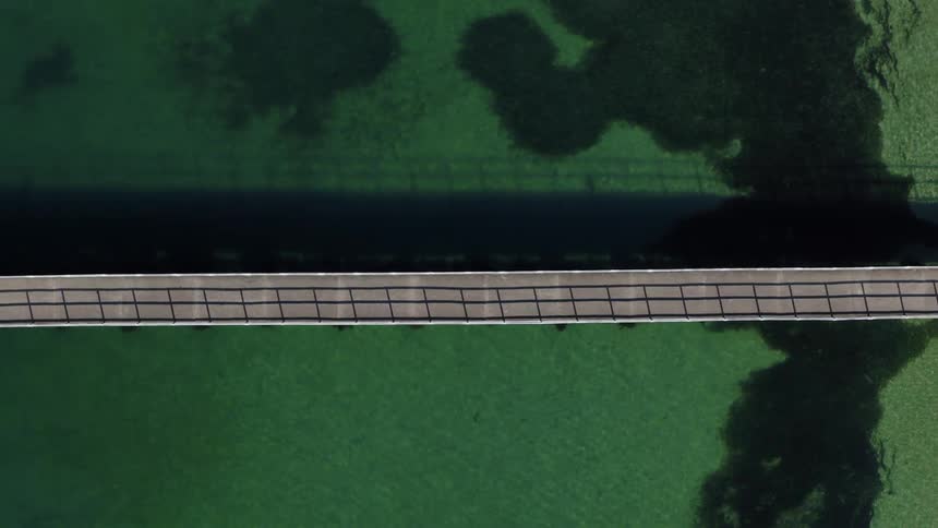 A static topdown aerial shot shows a straight bridge crossing over beautiful clear green water casting a dark shadow directly below it