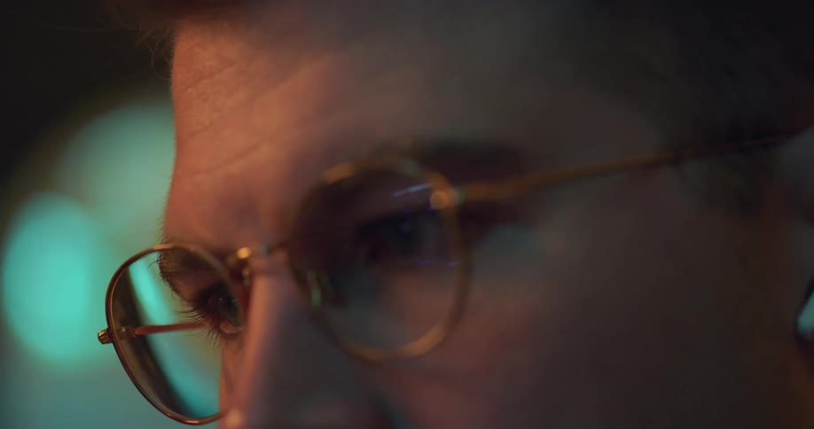 A person wearing glasses is looking at a computer or device in a dimly lit room with a blue light reflecting on their face the reflection of the screen is visible in their glasses as they blink and move their eyes