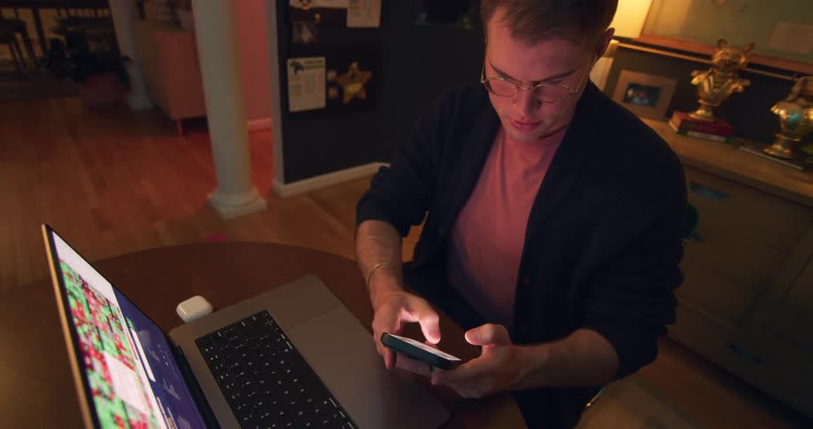 A focused man with glasses and earbuds is sitting at a desk looking at his laptop and phone he appears to be working or managing something on his devices in a dimly lit cozy room