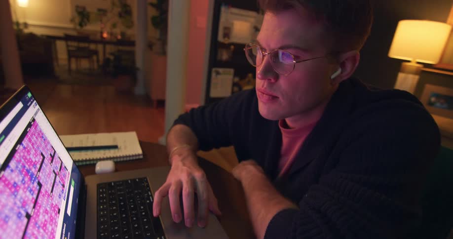 Man wearing glasses and earphones works on a laptop at a table with a notebook and lamp in the background