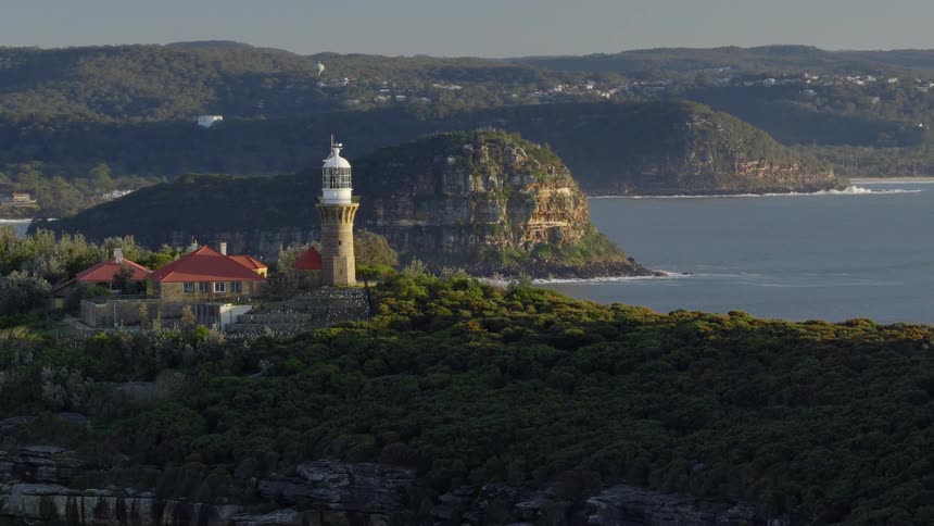 A scenic aerial shot sweeps past a historic lighthouse and keepers house perched on a lush green headland overlooking the ocean