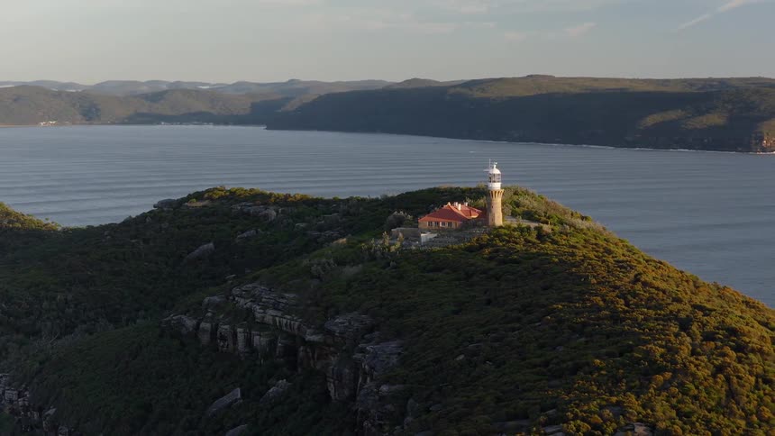 Lighthouse and buildings on a forested mountain along the coast with mountains across the river
