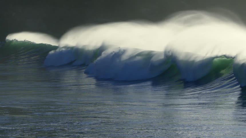 Large powerful waves are seen crashing in a continuous motion across the ocean with white foam spraying from their crests under a dark sky