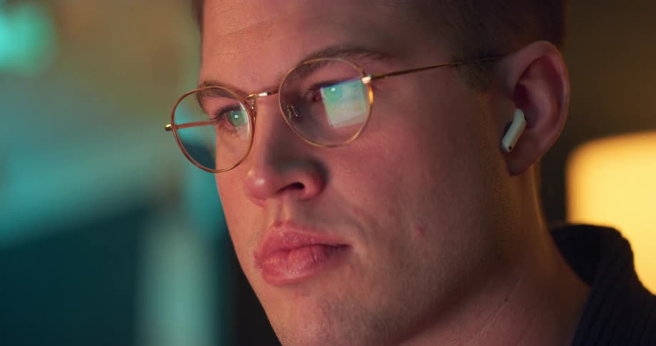 A close view shows a man with short brown hair wearing a white wireless earbud in his right ear and round gold glasses that reflect a blue light from a screen he is watching