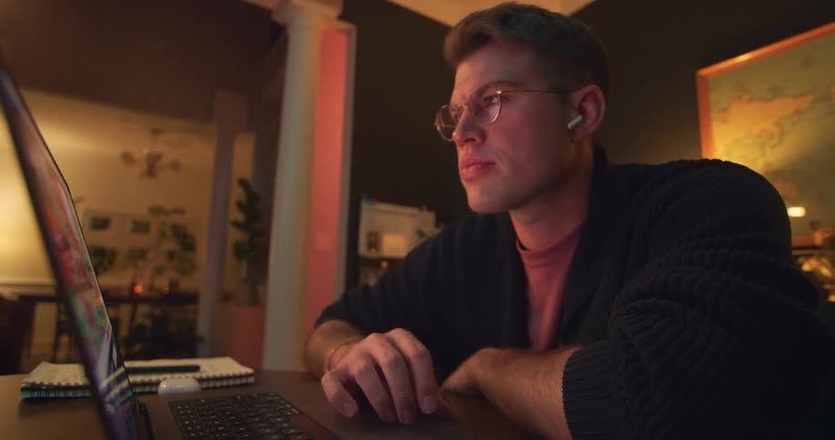 A man wearing glasses sits at a table while using his laptop computer