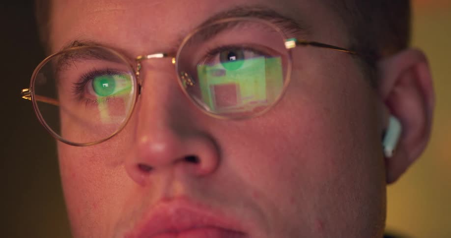 The face of a young man wearing reading glasses that is reflecting the image of a computer screen