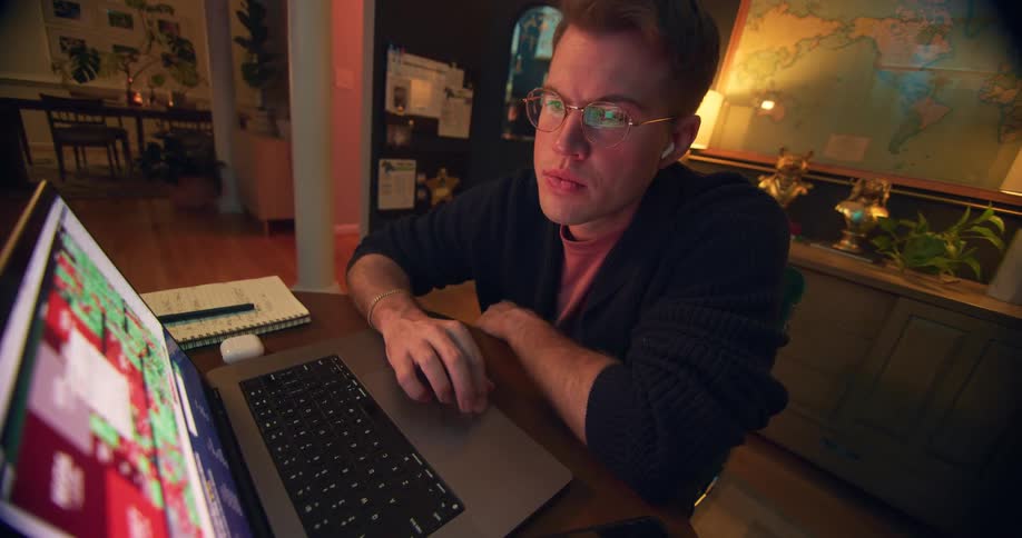 The video shows a man with short reddishblonde hair and glasses sitting at a desk hes wearing a dark cardigan and wireless earbuds and hes focused intently on the laptop screen in front of him