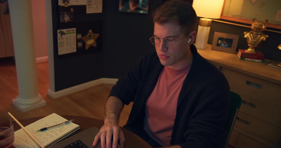 A young man wearing reading glasses and is sitting in front of a laptop and is studying writing something down in a notebook