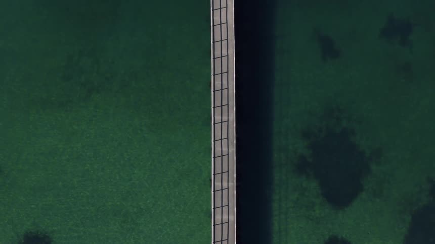 A slow topdown aerial shot pulls directly upwards from a long straight bridge that cuts vertically across calm dark green water