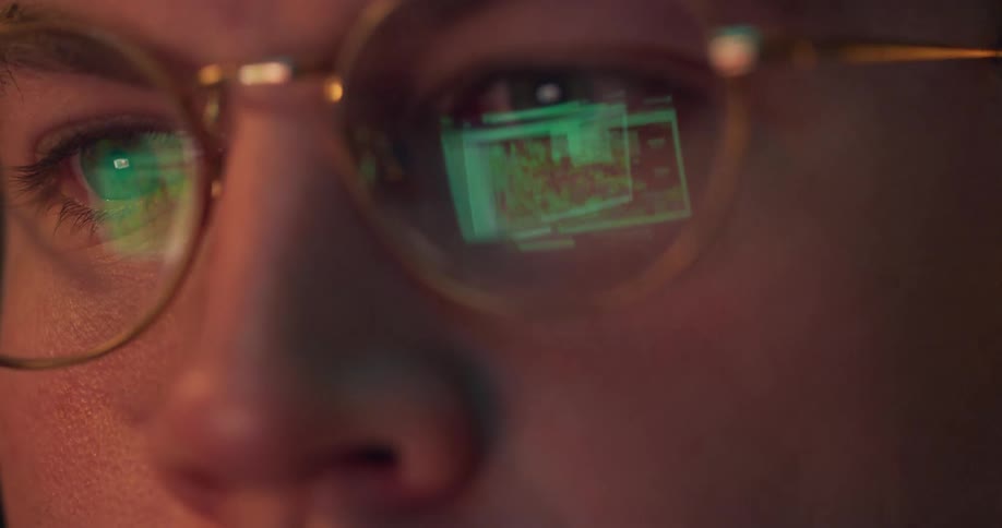 Monitor glow reflects in goldrimmed glasses tight on concentrating eyes